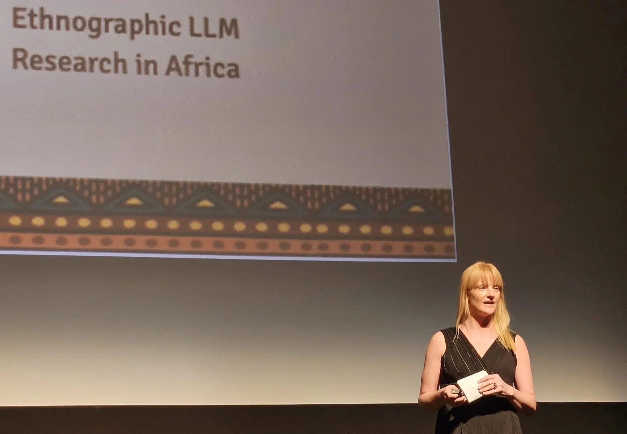 A woman with long blonde hair, wearing a sleeveless black dress, holding a notepad and speaking on stage in front of a large screen. The screen displays a presentation slide titled 'Ethnographic LLM Research in Africa' with a decorative border and pa