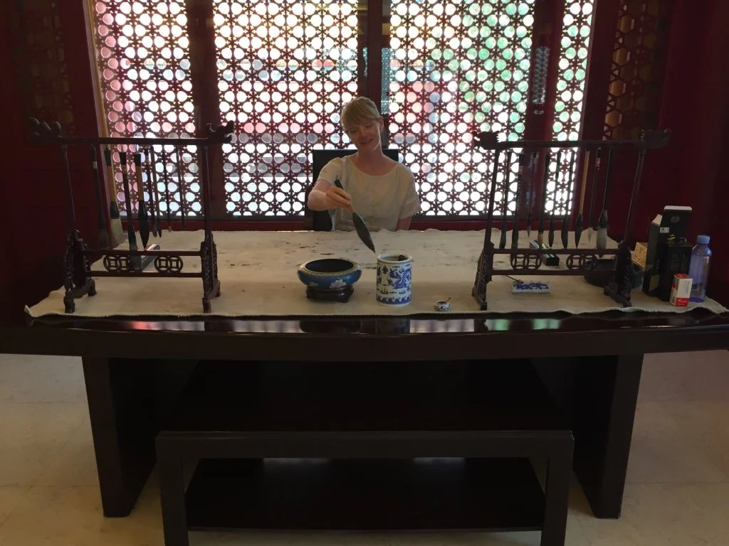 A woman sitting behind a wooden table, holding a spoon, with decorative wooden stands, a bowl, and a ceramic container on the table. The background features a richly patterned screen with geometric designs.