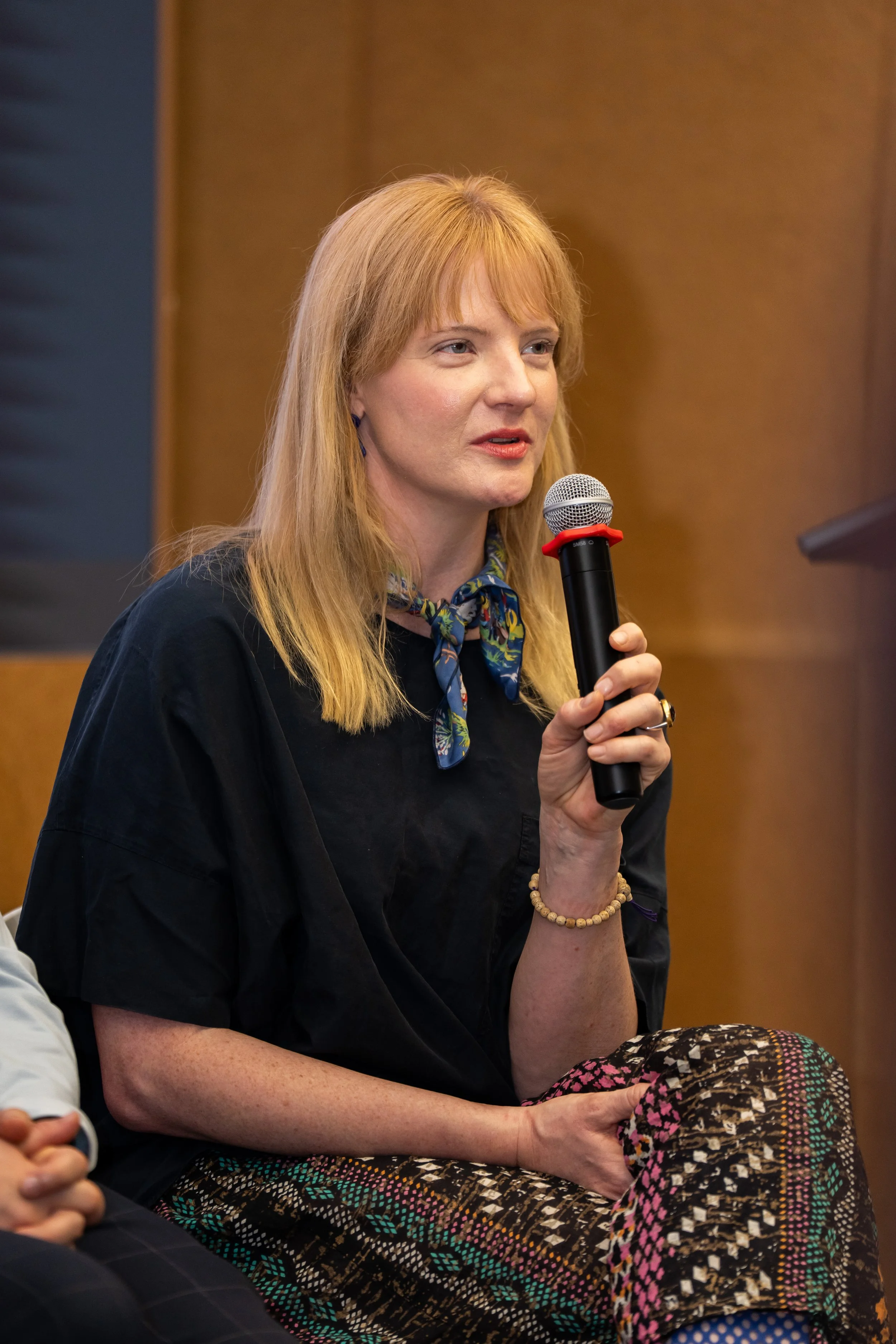 A woman with red hair, wearing a dark top and patterned pants, is holding a microphone and speaking during an event or discussion.