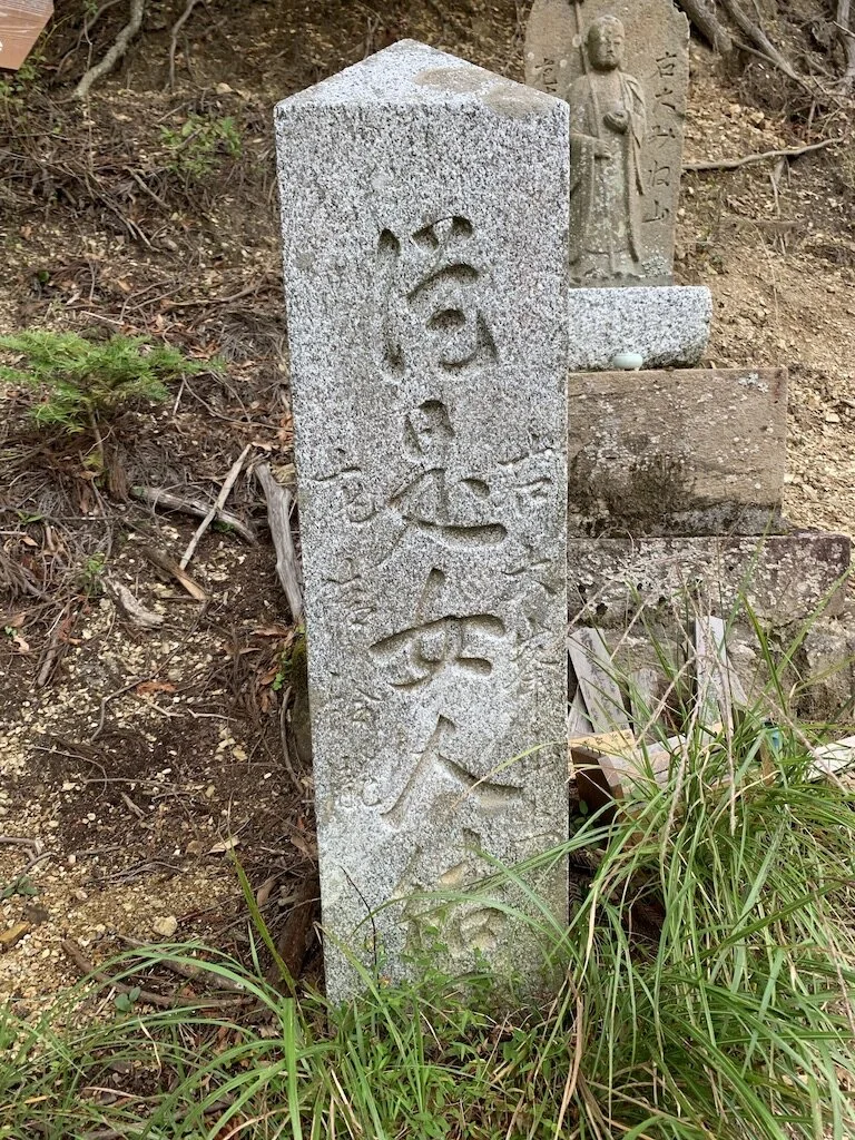 A vertical stone marker with Japanese characters engraved, situated outdoors among grass and dirt, with a small sculpture of a standing figure and additional stone markers in the background.