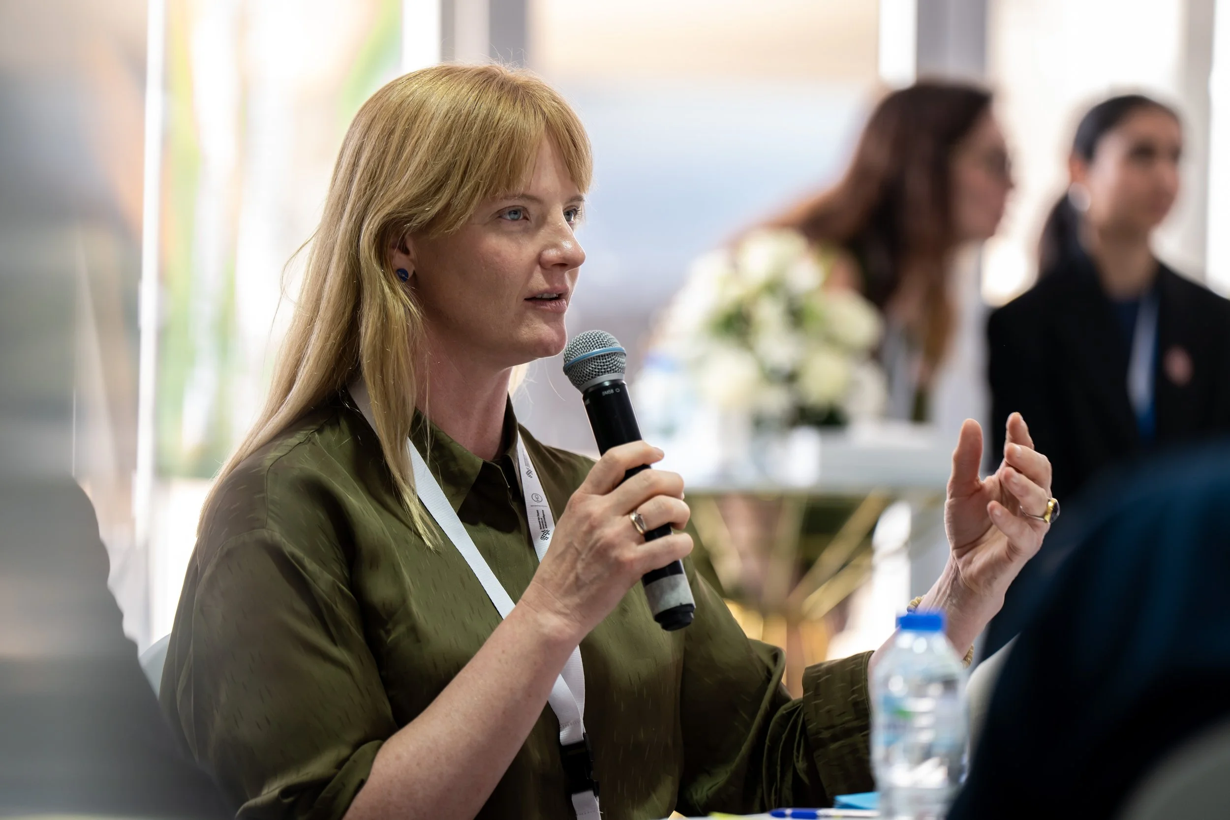 A woman with red hair and an olive green shirt speaking into a microphone during a meeting or conference, with blurred people and floral arrangements in the background.