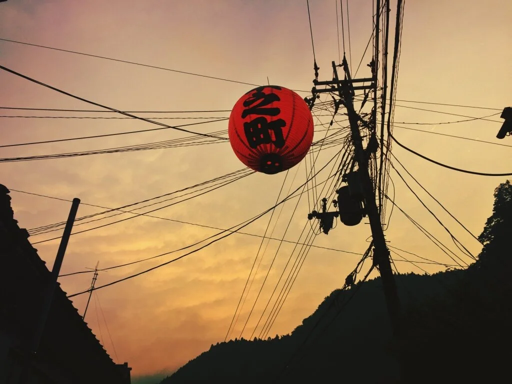 Red paper lantern hanging from overhead wires during sunset, with silhouetted power line poles and mountain in the background.