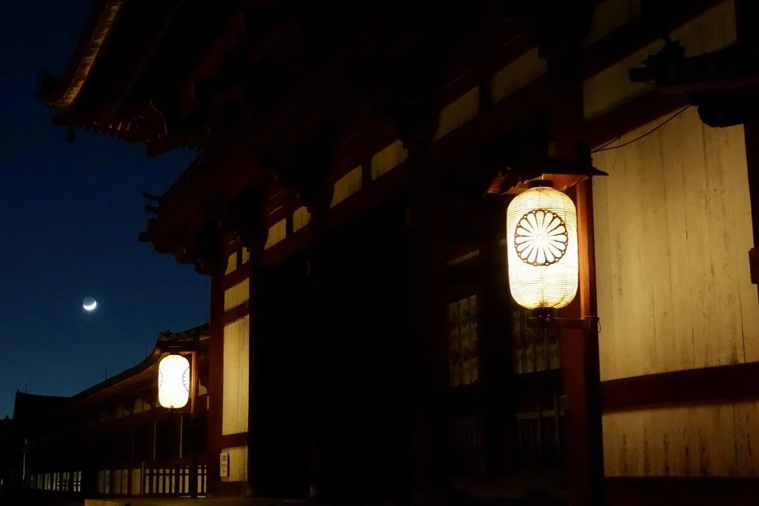 Nighttime photograph of traditional Japanese building with lanterns hanging outside, a crescent moon visible in the dark blue sky.