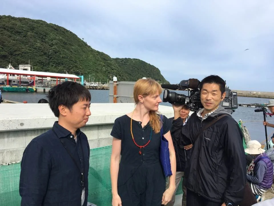 A group of people, including a man with a video camera, a woman with red hair and a blue dress, and a young man with black hair in a navy shirt, standing near a waterfront with boats and green hills in the background.