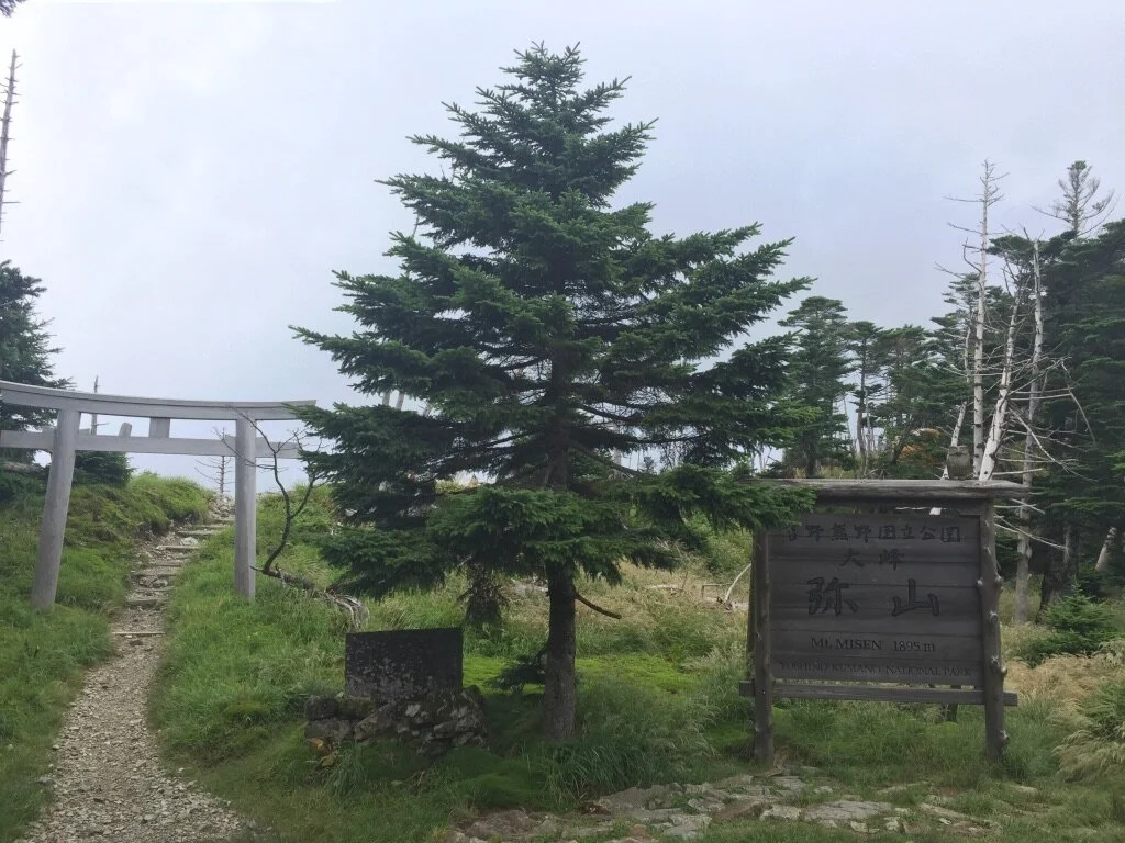 A dirt trail leading up to a Torii gate on a hillside with a pine tree, a wooden sign, and other trees in the background, indicating the summit of Mt. Misen at 1,895 meters.