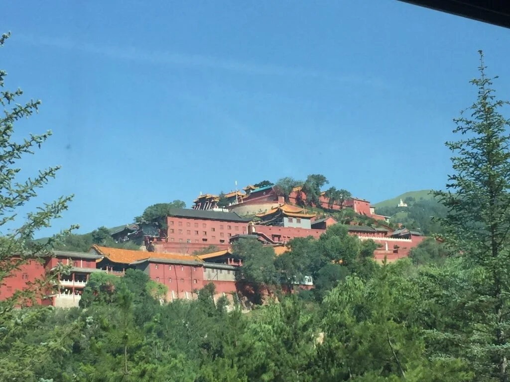 A large traditional Chinese-style palace or temple complex on a hill surrounded by trees and greenery, with a clear blue sky overhead.
