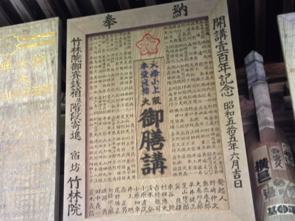 A framed Japanese document with vertical Japanese text, a red stamp, and a blue stamp, mounted on a wooden wall with other similar documents.