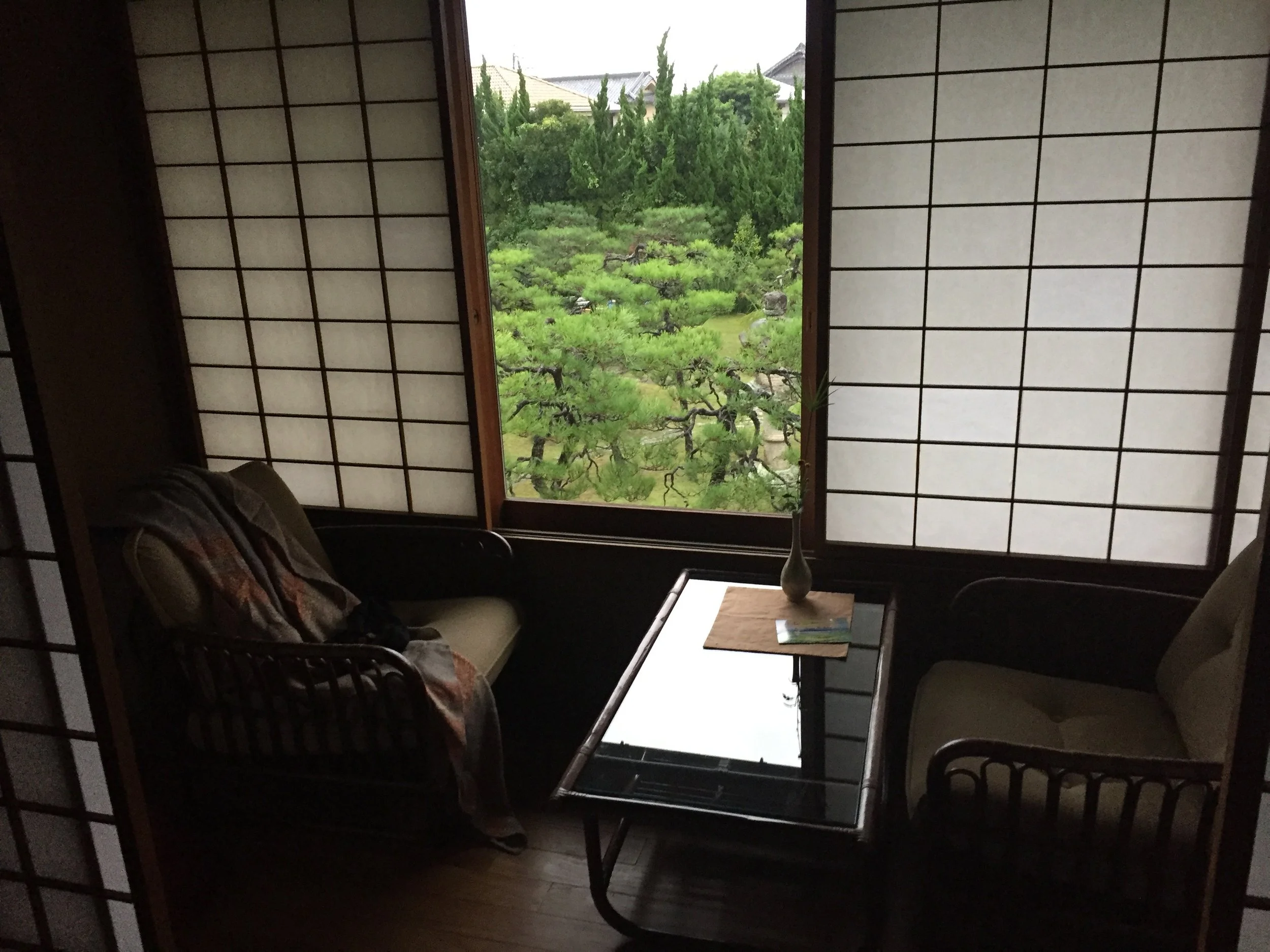 A cozy room with traditional Japanese shoji screens, a large window overlooking a lush green garden, a small table with a vase and a book, and two chairs with cushions, one of which has a blanket draped over it.
