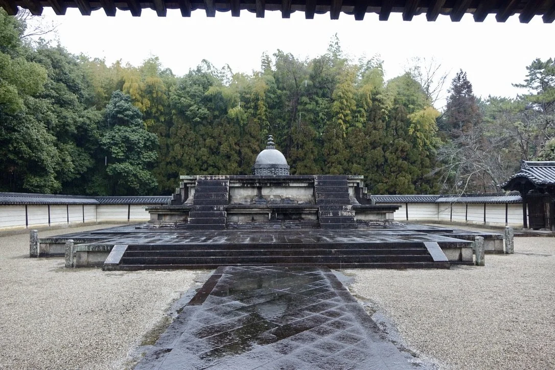 A Buddhist shrine with a large, black stone structure and a small dome on top, surrounded by a gravel courtyard and a white wall, in front of green trees.