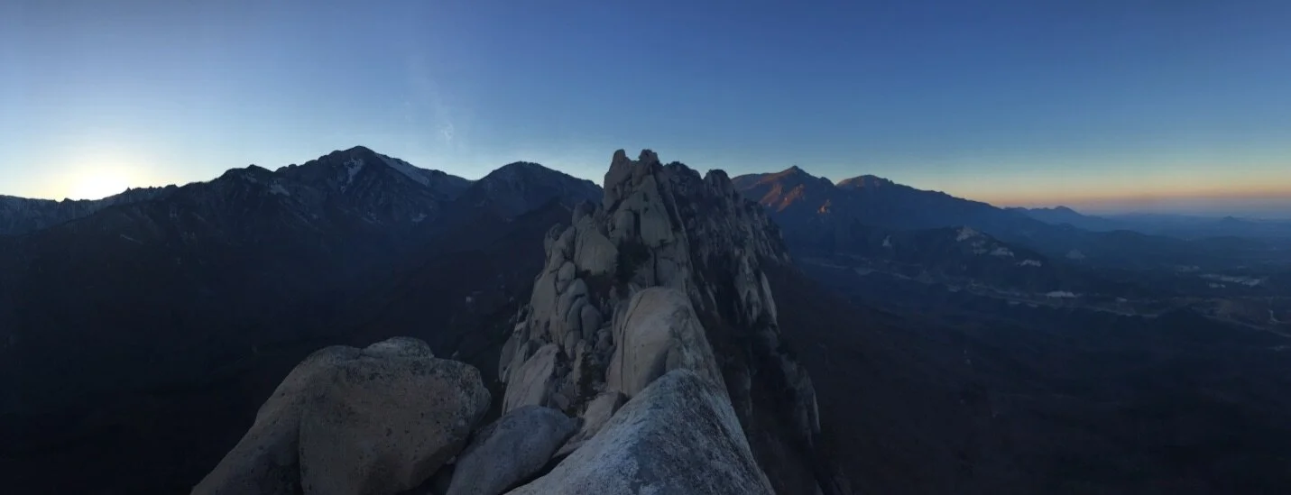 Panoramic view of rugged mountain peaks at sunrise with a rocky ridge in the foreground and a distant mountain range under a clear sky.