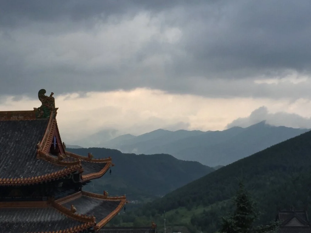 Traditional Asian temple with ornate rooftops against a backdrop of misty mountains and cloudy sky.