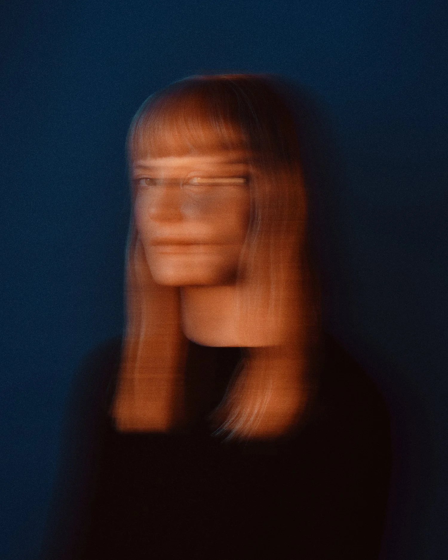 A portrait of a person with red hair against a dark blue background, with a motion blur effect distorting their face and hair.