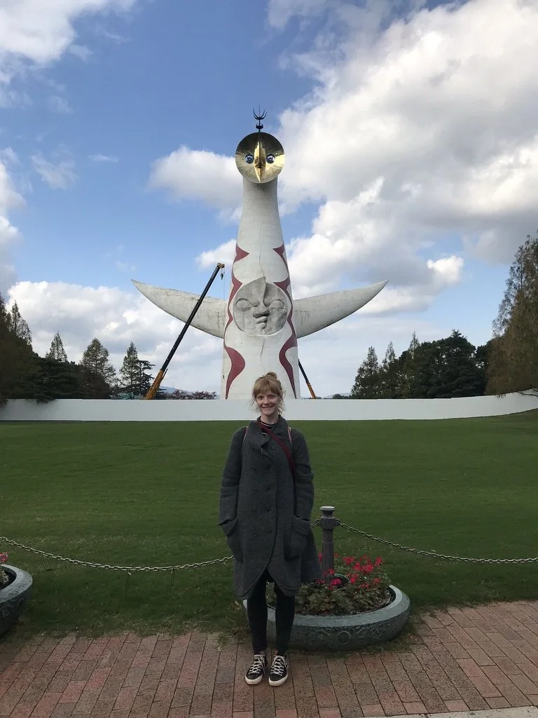A smiling young woman standing in front of the Tower of the Sun sculpture in a park, with green grass, flowers, and trees in the background.