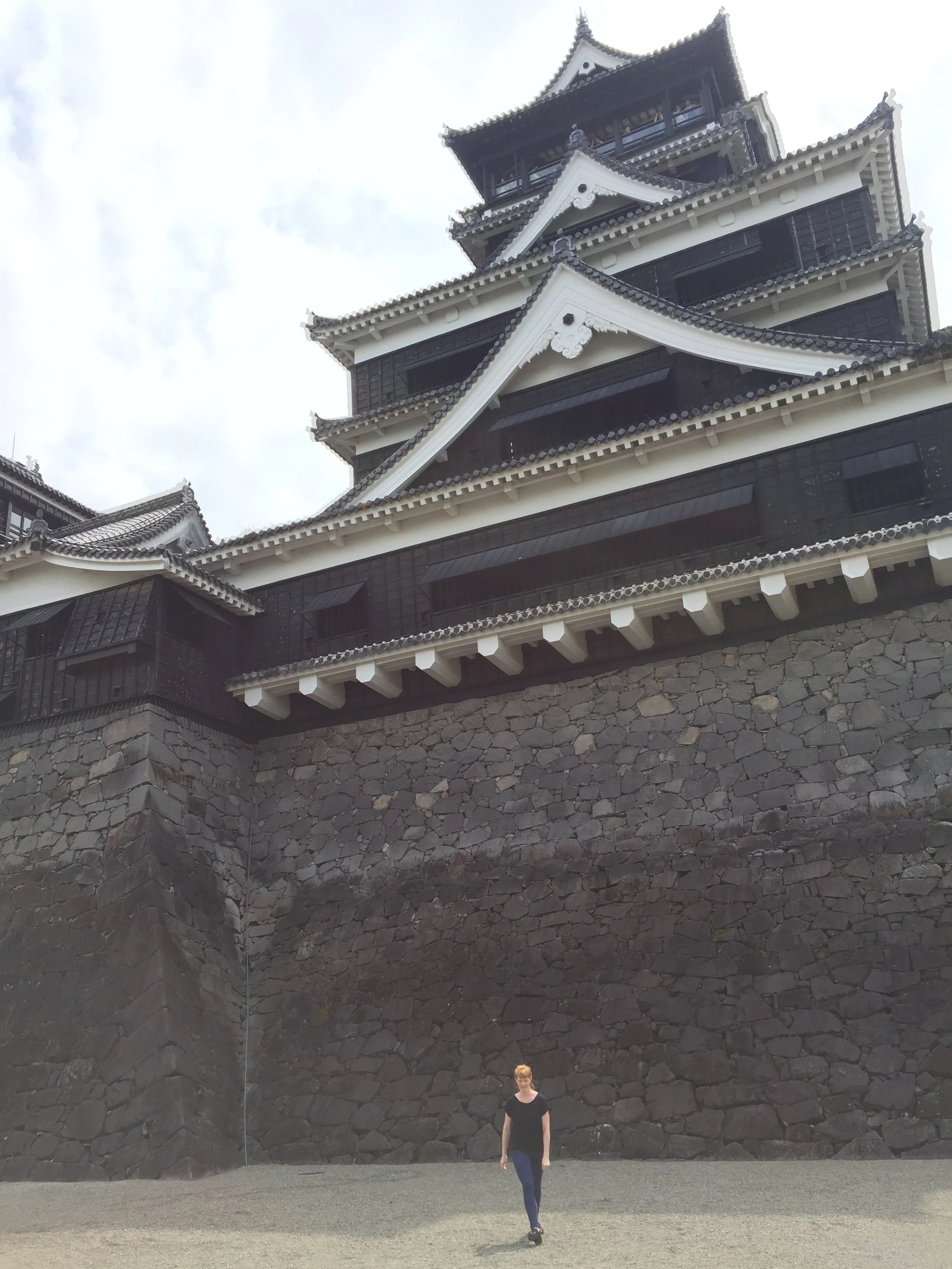 A person walking in front of a traditional Japanese castle with dark black walls, white detailing, multiple tiers, eaves, and a stone foundation.