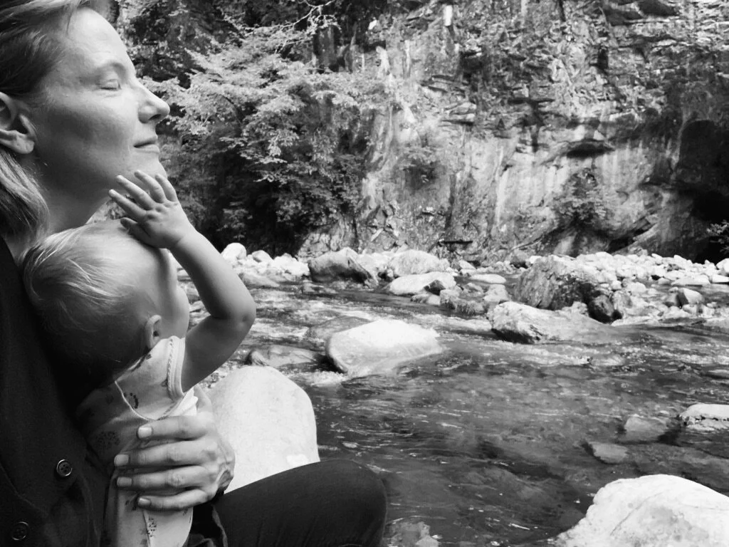 A woman with closed eyes, holding a young child near a river with rocks and a rocky cliff in the background.