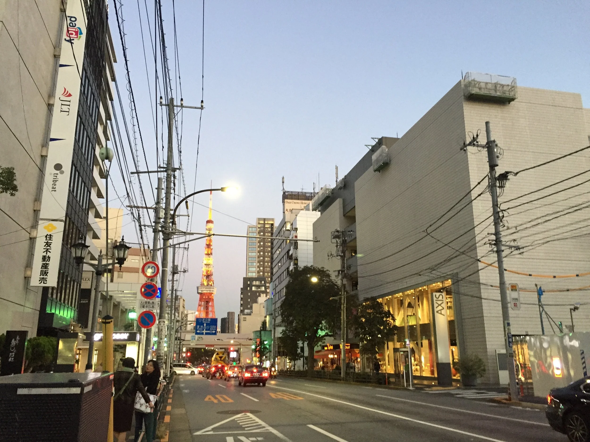 Evening street scene in Tokyo with mid-rise buildings, traffic, overhead power lines, and Tokyo Tower visible in the distance.