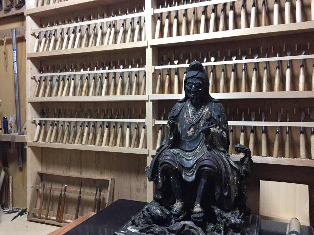 A black wooden statue of a seated figure in a workshop with shelves filled with chisels and carving tools in the background.