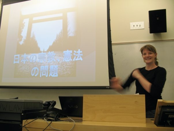 A woman standing next to a projection screen in a classroom, presenting a slide with Japanese text and an image of a torii gate, with laptop and equipment on the desk in front.