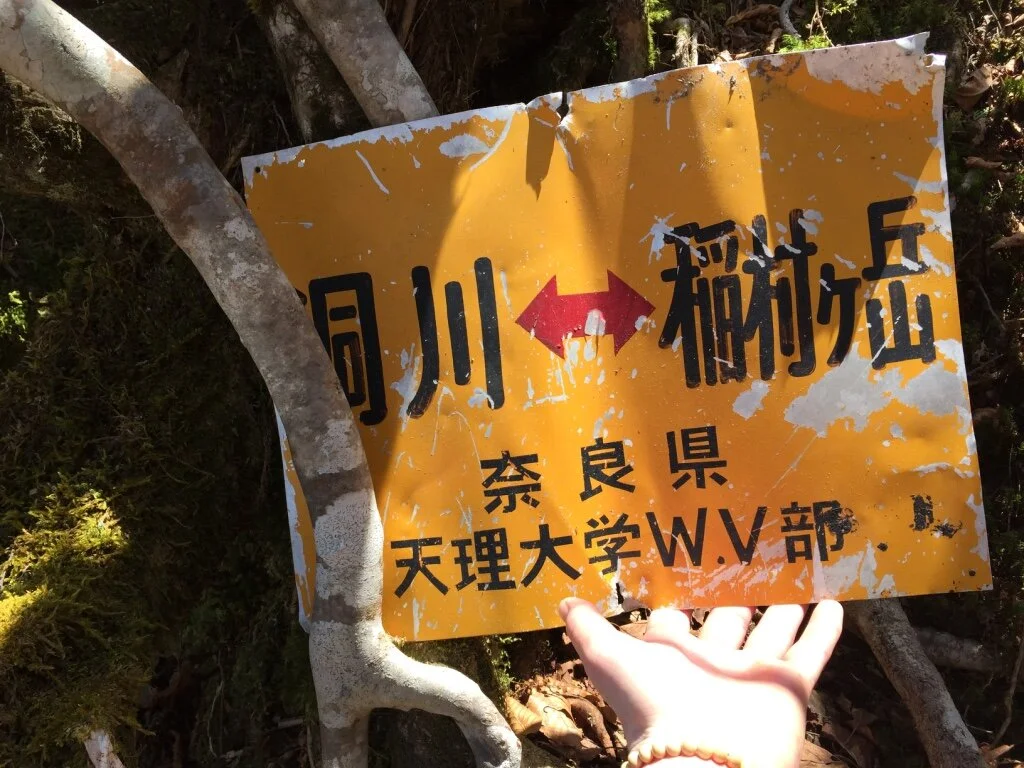 Worn yellow outdoor sign in Japanese with black and red text, partially obscured by a person's hand, surrounded by trees and moss.