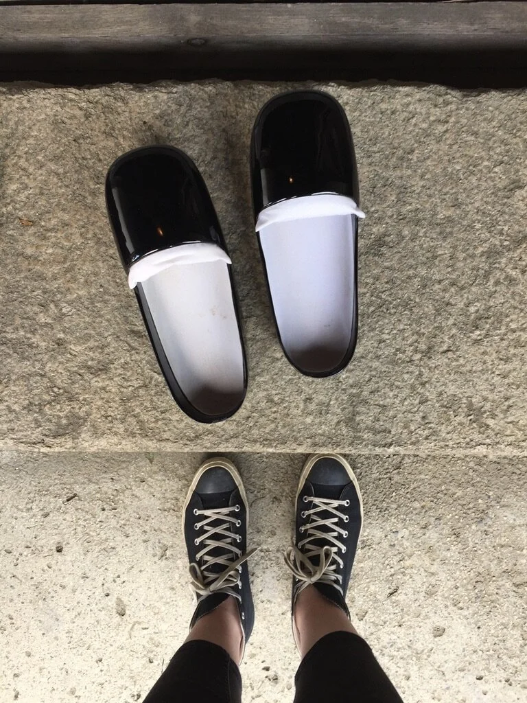 A pair of black slip-on shoes with white socks and laces, seen from above, standing on a concrete surface with a pair of black shoes on top of the person's shoes.