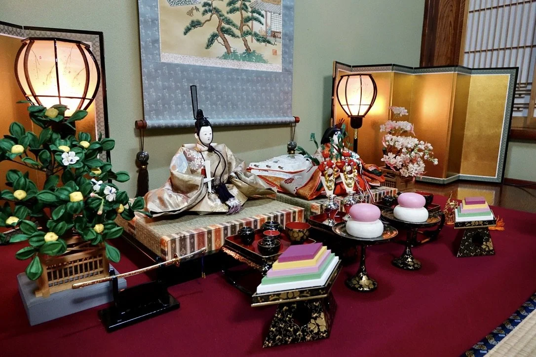 Traditional Japanese display with a Hina doll, lanterns, cherry blossom decorations, colorful paper stacks, and ceremonial items on a red carpeted surface, set against a room with a gold folding screen and a wall hanging with a tree painting.