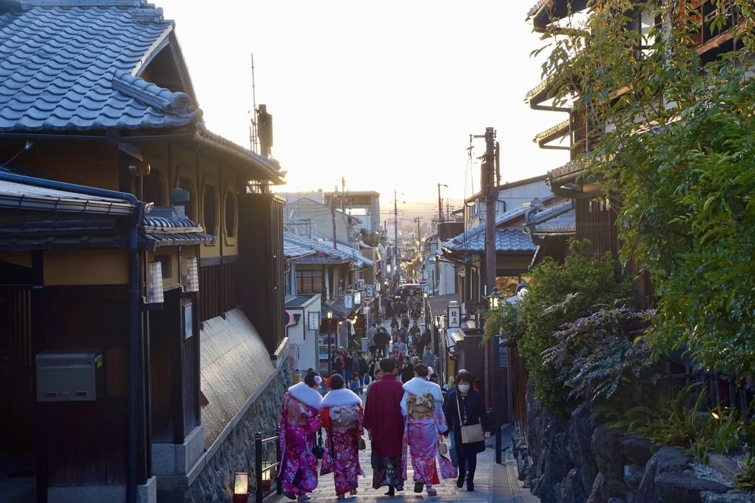 A street scene in Japan at sunset with people walking, some in traditional kimonos, surrounded by traditional Japanese buildings with tiled roofs and greenery.