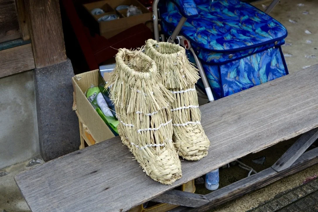 A pair of straw slippers placed on a wooden surface, with a blue patterned bag and a cardboard box in the background.