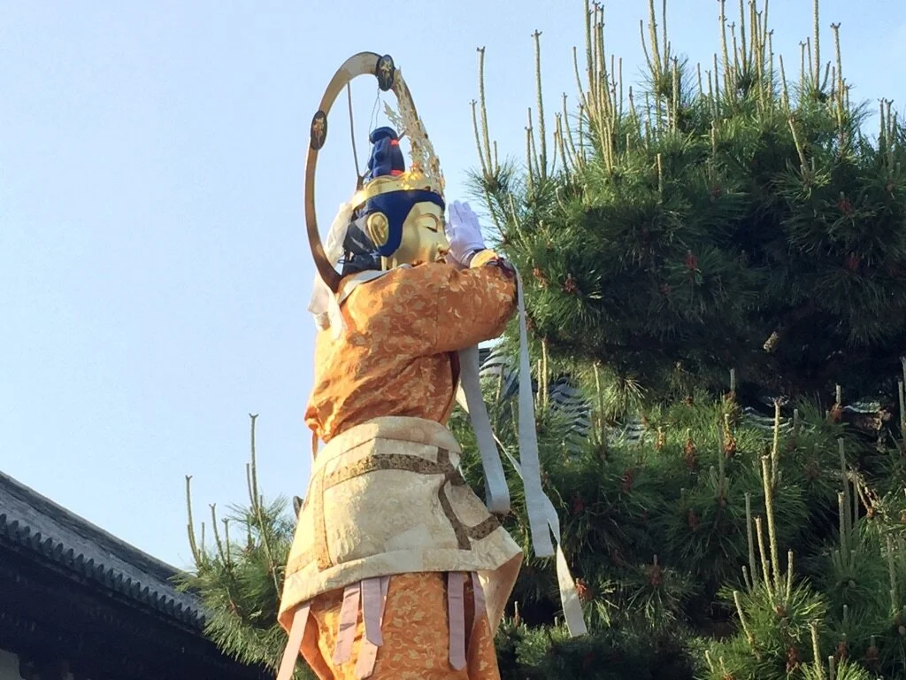 A traditional Japanese or Chinese parade float featuring a large, ornate figure of a person with a headpiece, wearing colorful robes, and holding a staff or weapon, standing next to a pine tree.