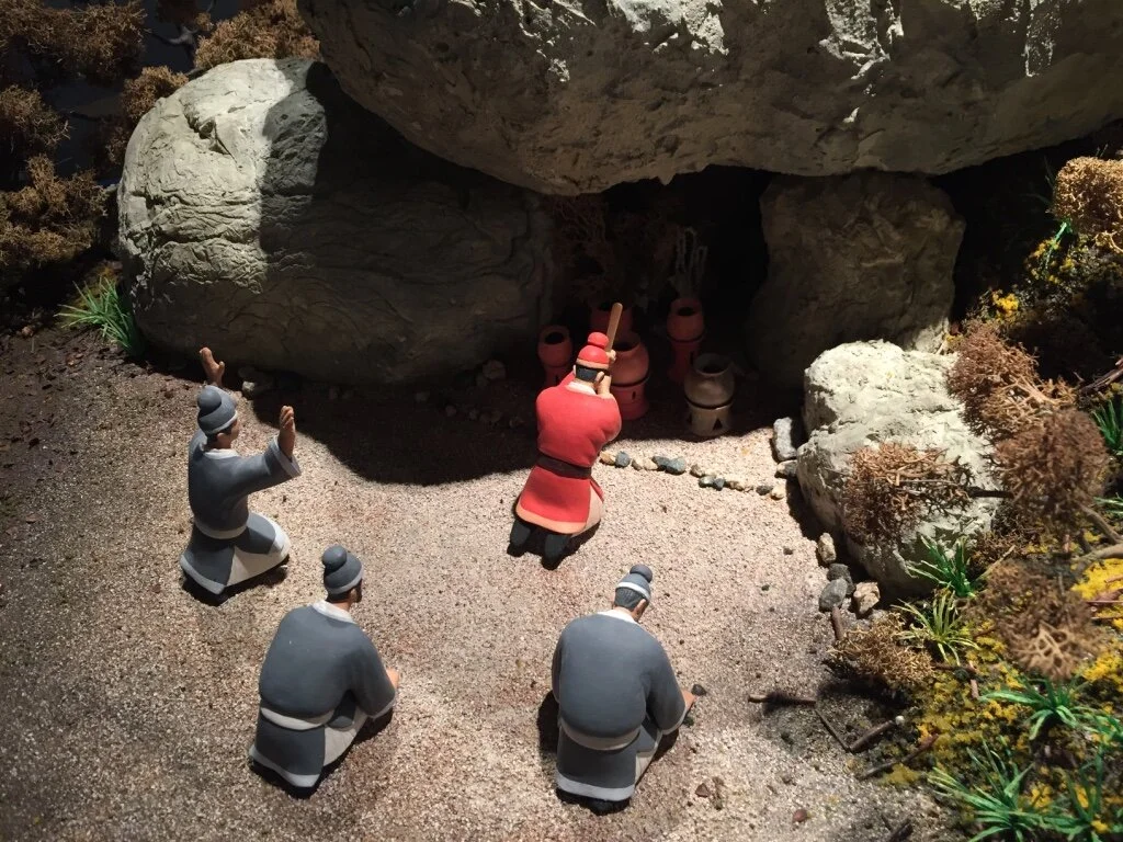 Miniature figures of people in robes kneeling and praying in front of a cave with several clay pots and a figure of a person in a red robe and robe, surrounded by rocks and plants.