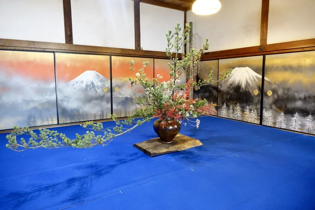 A traditional Japanese room with a blue carpet, a flower arrangement in a brown vase on a wooden platform, and a backdrop of painted panels depicting Mt. Fuji and nature scenes.