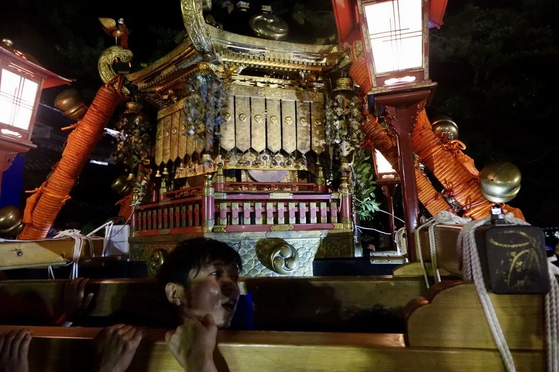 A traditional Japanese festival float, or mikoshi, decorated with gold, red, and black accents, large orange rags, lanterns, and gold ornaments, carried during a festival at night.