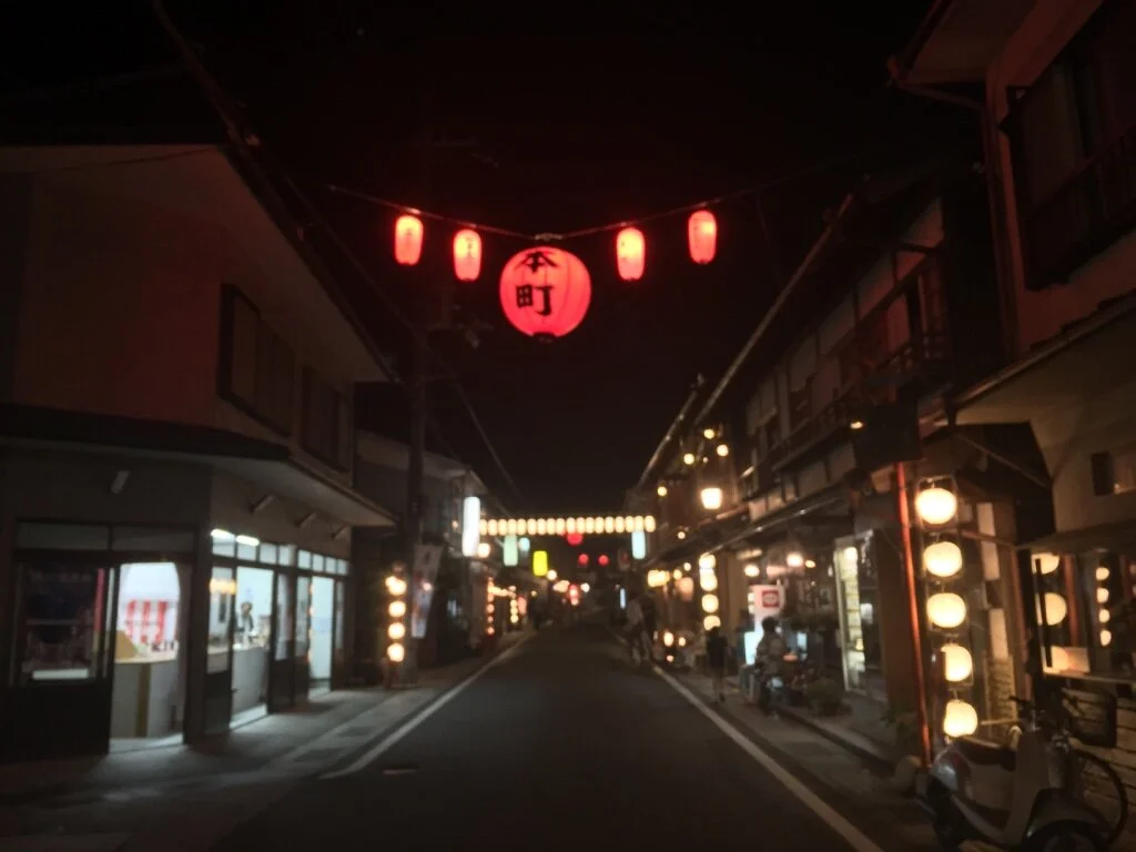 A nighttime street scene in Japan with red paper lanterns hanging overhead, illuminated shop fronts, and a narrow road leading into the distance.