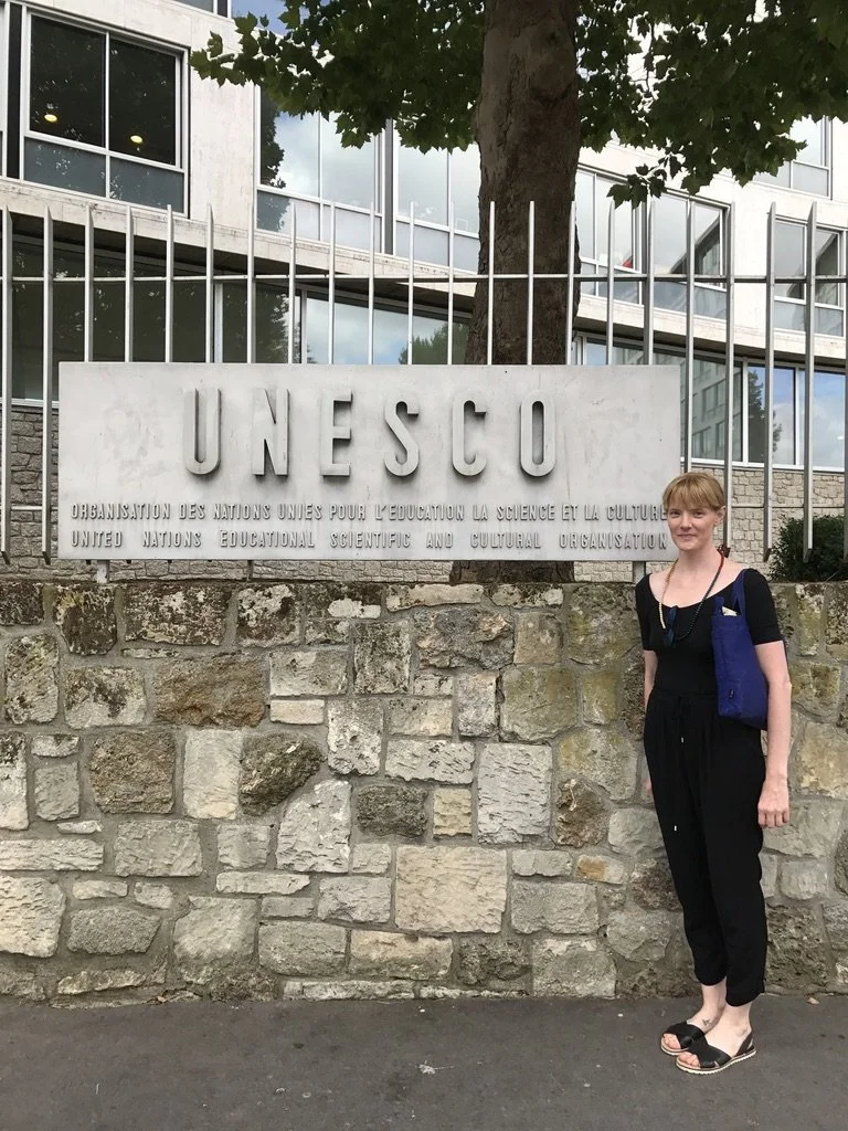 A woman standing next to a UNESCO sign that reads 'UNESCO Organisation des Nations Unies pour l'éducation, la science et la culture / United Nations Educational, Scientific and Cultural Organisation' in front of a stone wall and a building with large