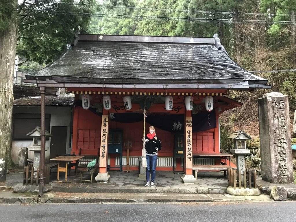 A woman standing in front of a small traditional Japanese shrine with a rope and bell. The shrine is red with hanging lanterns and is surrounded by trees.