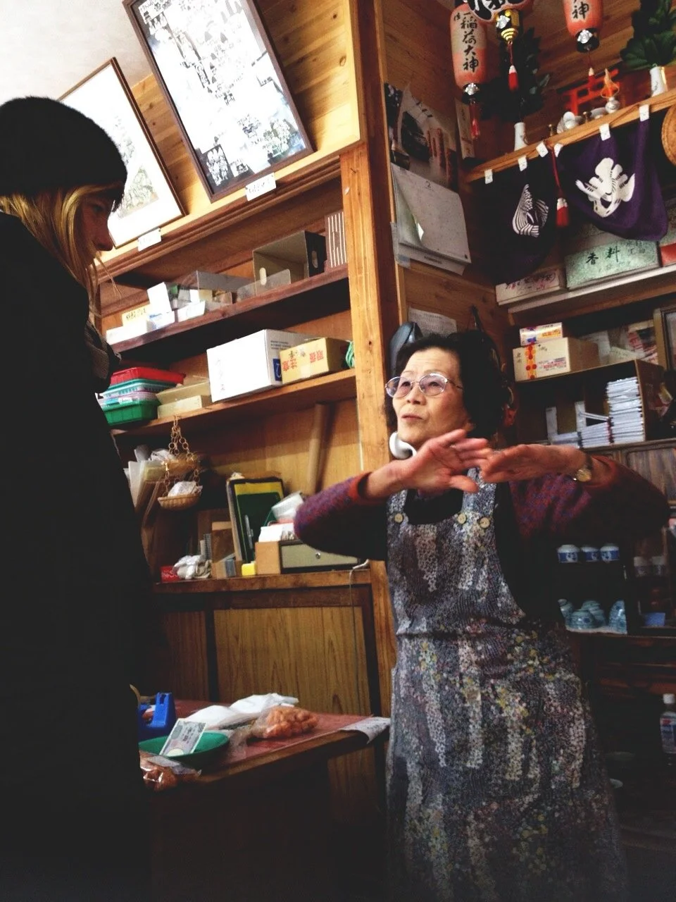 An elderly woman wearing glasses and an apron is gesturing with her hands while talking to a younger woman in a black coat inside a store with wooden shelves, books, and Japanese decorations.