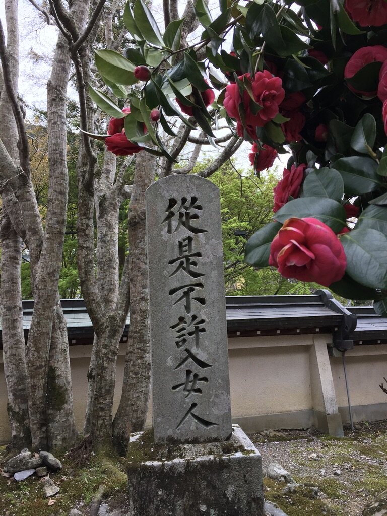 Stone monument with Japanese characters engraved, surrounded by trees and pink flowers, in a garden or outdoor setting.
