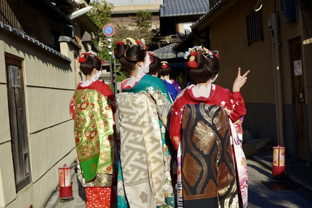 Group of women dressed as Geishas in traditional kimonos and elaborate hairstyles walking down a street with wooden houses in Kyoto, Japan.