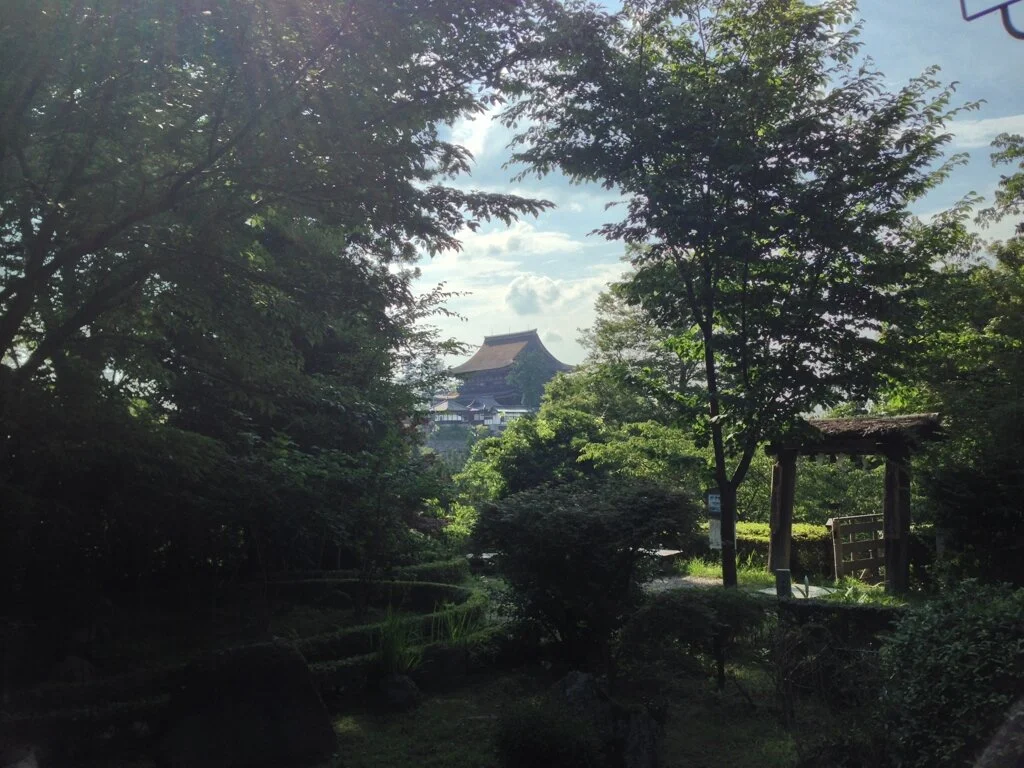 A lush garden with various trees and shrubs, a small wooden gate on the right, and a traditional building in the distance under a partly cloudy sky.