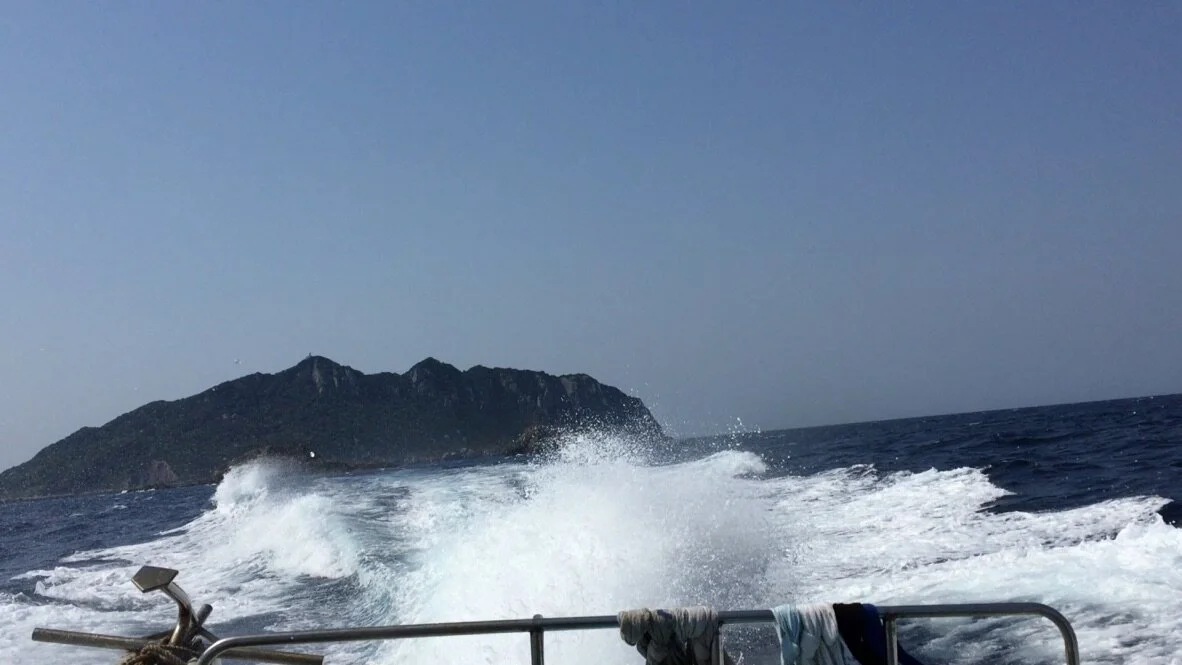 View from a boat moving through the ocean, with a small island in the distance and waves splashing behind the boat.