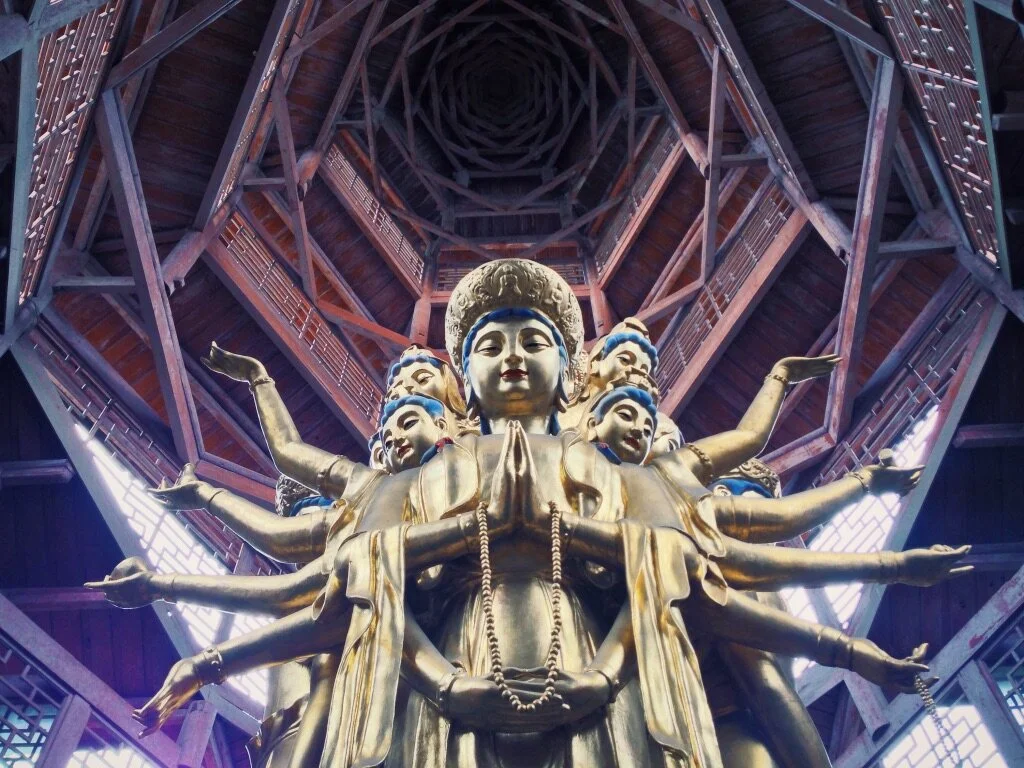 A large golden multi-armed statue of a deity with a serene face inside a wooden, geometric, octagonal structure with spiral design above.