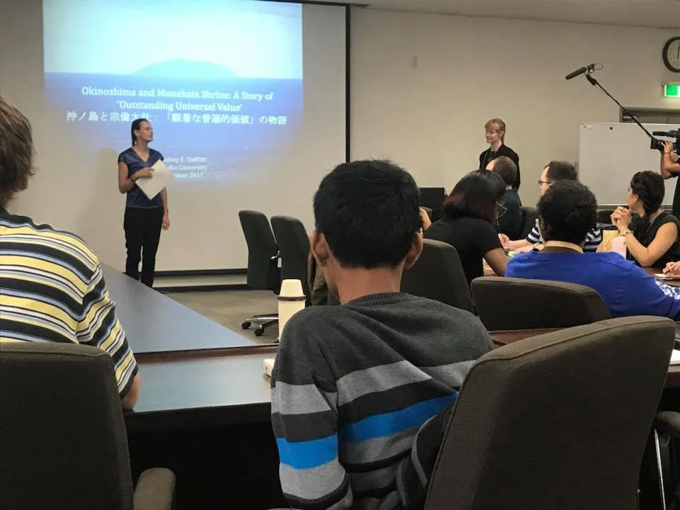 A woman stands at the front of a conference room giving a presentation on a large screen, with seated audience members watching. The presentation slide is titled "Okinoshima and Munakata Shrine: A Story of 'Outstanding Universal Value'".