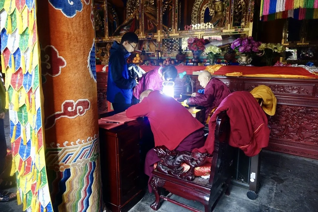 Buddhist monks and laypeople praying and making offerings inside a temple with ornate decorations and colorful flowers.