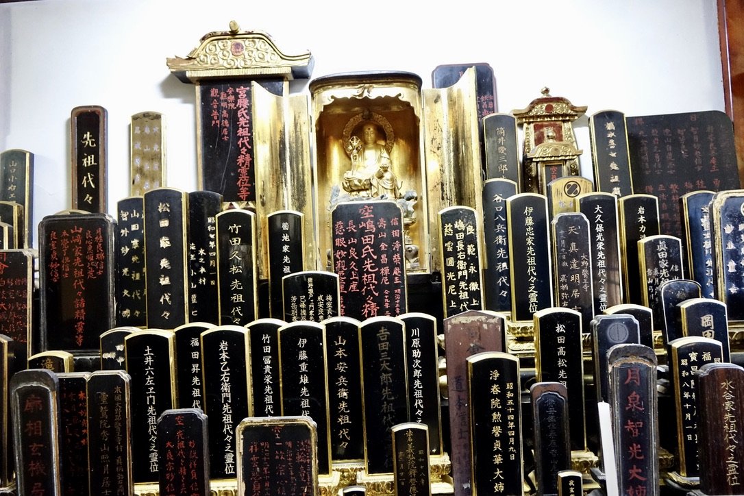 Collection of black and gold tombstones with red and gold Japanese inscriptions surrounding a small altar with a golden statue of a woman, in a shrine.