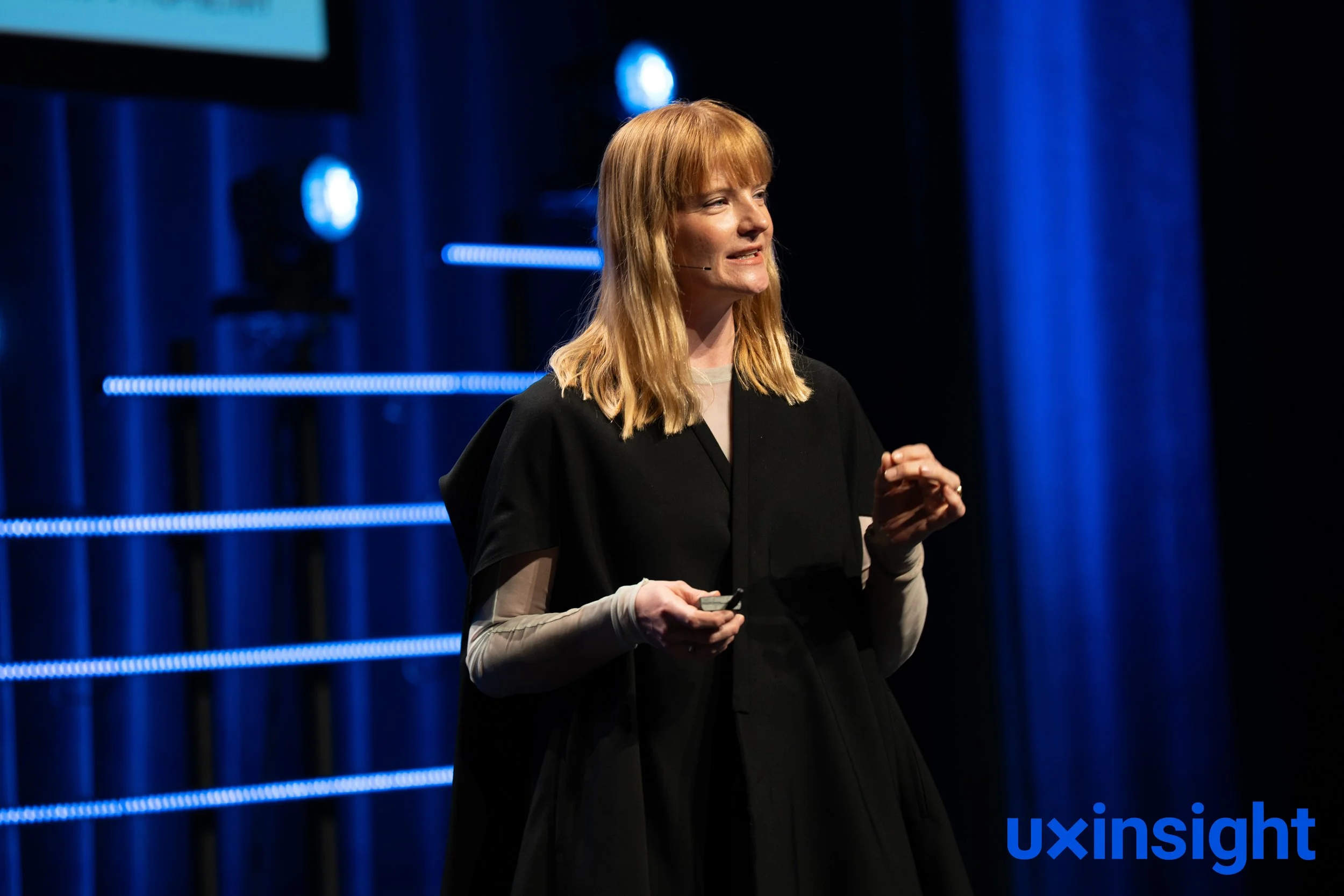 A woman with red hair wearing a black top and beige sleeves delivers a presentation on stage with blue lighting in the background.