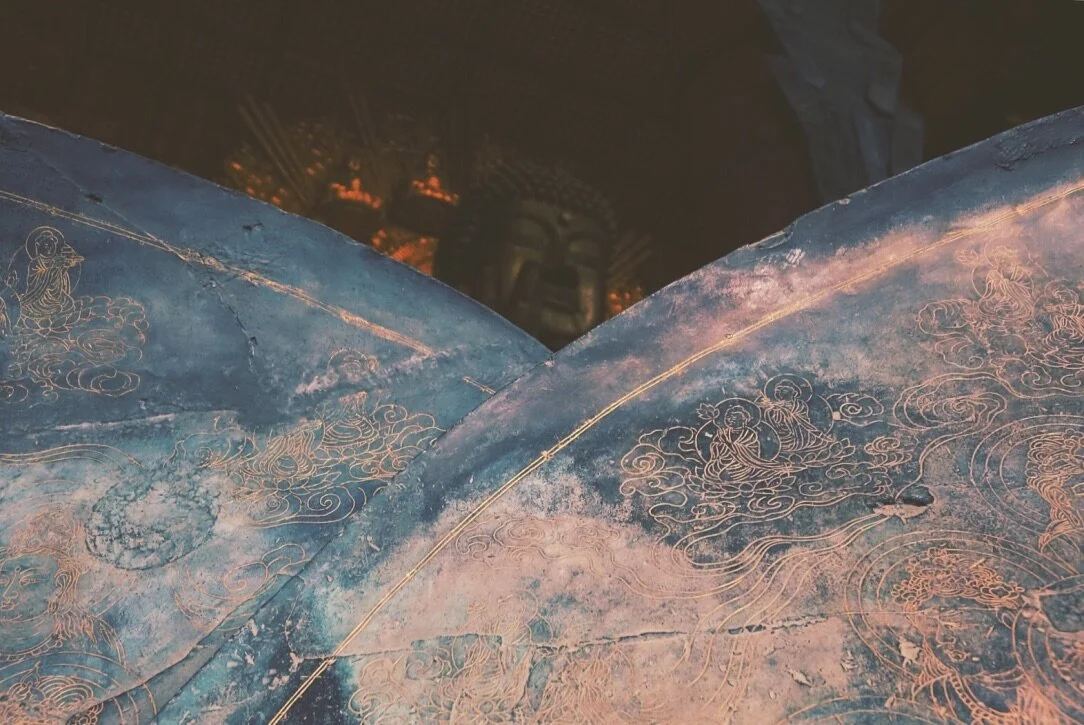 Close-up of an ancient, weathered blue and gold painted Buddhist parasol with intricate line art depicting Buddhist figures and clouds. In the background, a dark Buddha statue is partially visible.