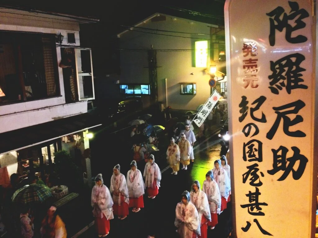 Nighttime street scene with a procession of people in traditional Japanese attire, including white coats and red skirts, walking along the street. Some people are holding umbrellas, and there are illuminated signs and buildings in the background.