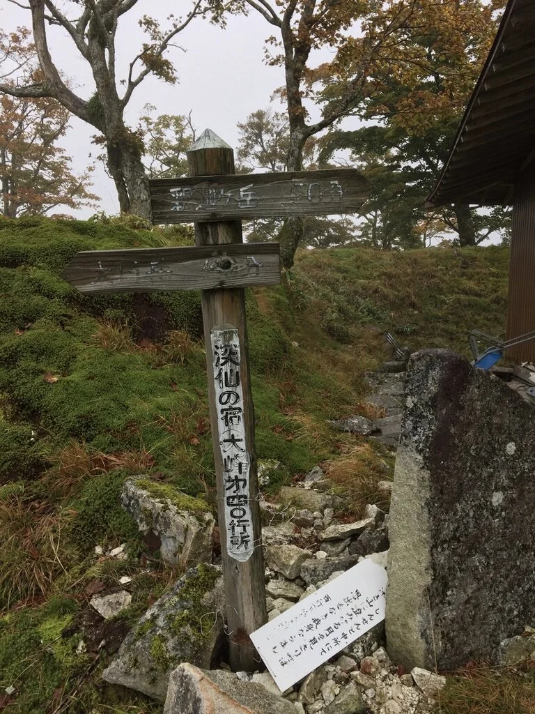 Wooden signpost on a rocky, moss-covered hillside with Japanese writing, surrounded by trees and foggy weather, next to a small building with a stone marker nearby.