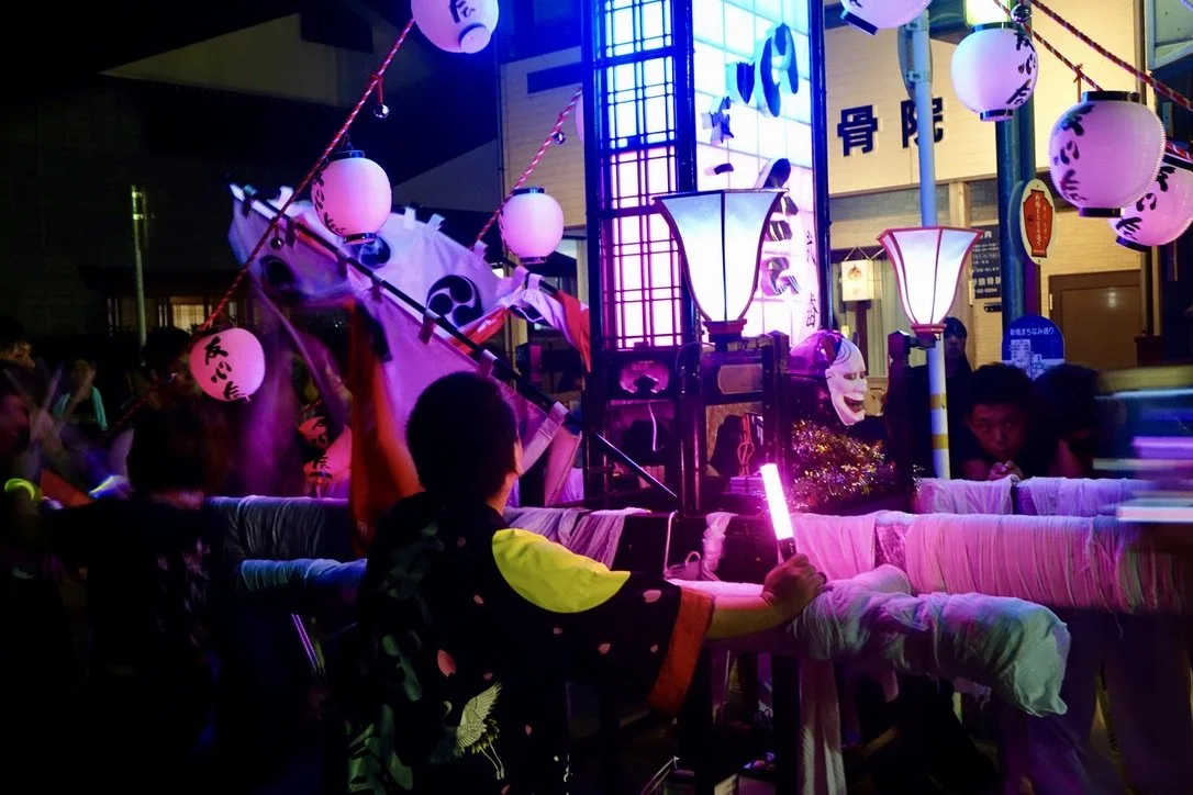 Nighttime scene of a traditional Japanese festival float decorated with paper lanterns, lanterns, and fabric, with people surrounding it, some holding lights, and a large illuminated sign in the background.