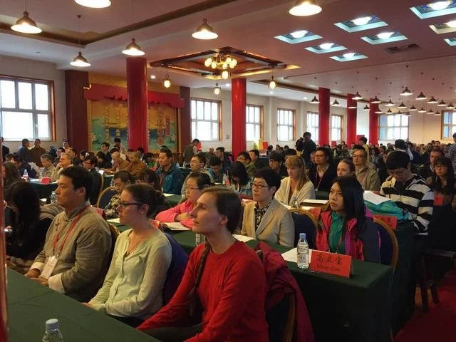 Audience seated in rows at a conference or seminar in a decorated hall with red columns and windows, wearing formal and casual attire.
