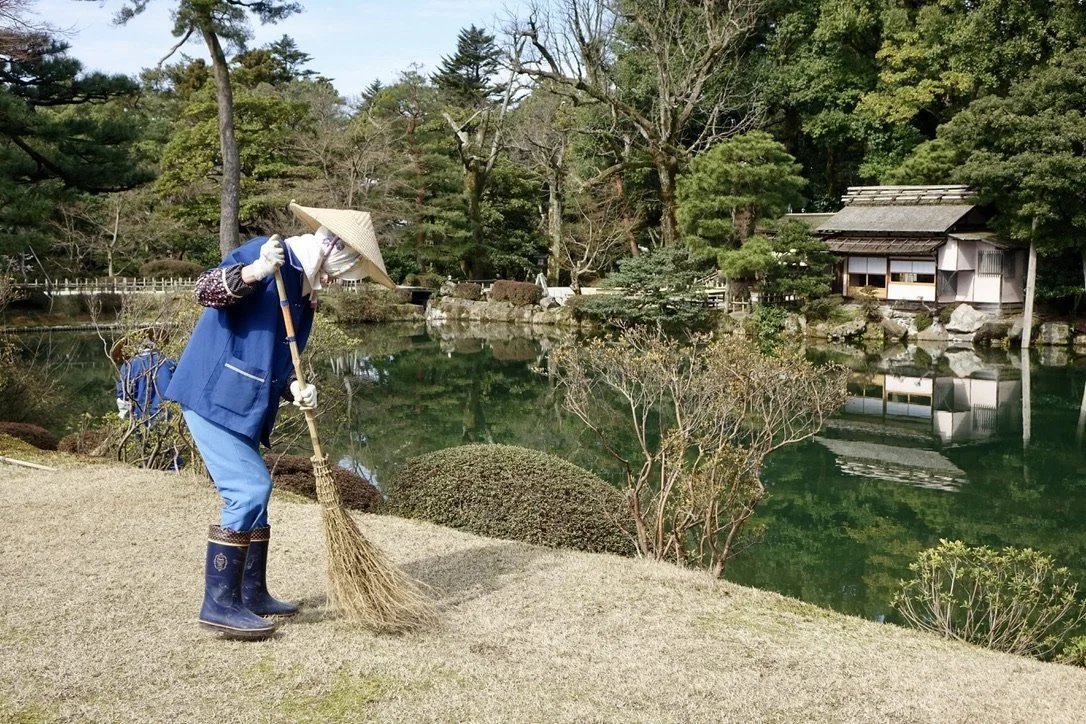A person wearing a blue coat, hat, and rain boots sweeping the ground near a pond with a traditional building and trees in the background.